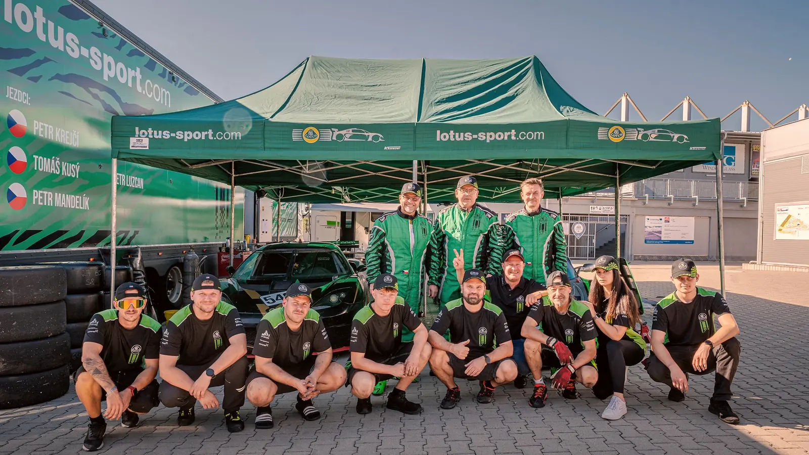 Tým KT Motorsport před stanem a kamionem v depu, jezdci v kombinézách, Evora 540 vzadu